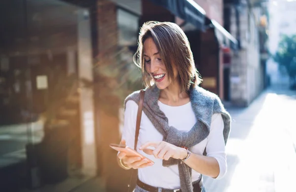 明るい日差しと携帯電話でのメッセージの下で通りに一人で立ってカジュアルな服で楽観的な笑顔の中年女性 — ストック写真