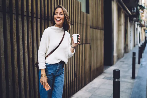 カジュアルな服装でスマートフォンを持つ中年女性を自信を持って笑顔と紙コップでコーヒーを飲みながら 現代的な建物の外観に対して路上でリラックス — ストック写真