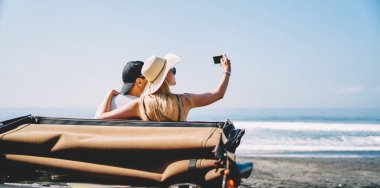 Arabanın yanındaki şapkalı genç bir çiftin yaz tatilinde deniz kenarında selfie çekerken görüntüsü.