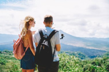 Tanınmayan genç turistlerin arka manzarası. Kadın ve erkek tepenin üstünde durup güneşli yaz gününde nefes kesici manzaralara hayranlık duyuyorlar. Günlük giysiler ve sırt çantaları giyiyorlar.
