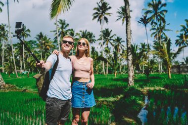 Güneş gözlüklü gülümseyen erkek turist, yaz günü egzotik ormanların yanında dururken kameraya selfie çekiyor ve sakin kız arkadaşına sarılıyor.