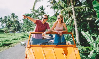 Genç bir adam ve bir kadın açık arabada güneşli bir yaz gününün tadını çıkarırken güneş gözlüğü ve günlük kıyafetler takıp gözlerini kaçırıyorlar.