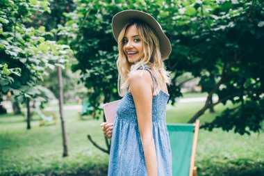 Şehir parkında gündüz vakti poz veren neşeli bir kız öğrencinin yarım boy portresi, şık şapkalı mutlu beyaz kız kişisel planlayıcısını tutuyor ve kameraya gülümsüyor.