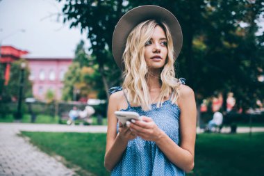 Cep telefonu cihazıyla tefekkür eden düşünceli bir kadın blogcu yayın metnini paylaşmayı düşünüyor, modaya uygun şapkalı dalgın hippi kız modern akıllı telefon cihazını tutuyor ve düşünüyor.