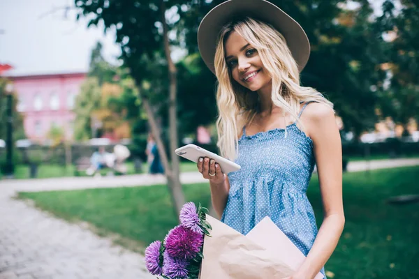 Bir sürü güzel yaz çiçekleri ve akıllı telefon cihazıyla neşeli hippi kız. Hafta sonu parkta poz veriyor. Şapkalı ve cep telefonlu neşeli beyaz kadın portresi.