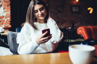Tahta masada oturan, boynuna bir fincan kahve değmiş ve cep telefonuyla sohbet eden sıradan bir kadın.