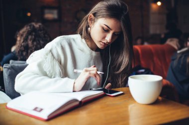Konsantre olmuş, günlük kıyafetli bir kız öğrenci. Üniversite için çalışırken ders kitabı ve cep telefonuyla masada oturuyor.