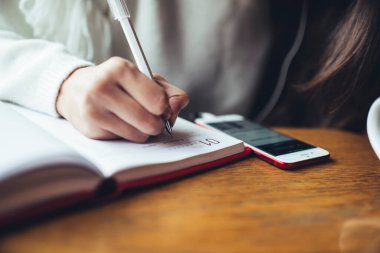 Tanımlanamayan kadın serbest çalışan, ahşap masada oturan, cep telefonu kullanan ve proje üzerinde çalışırken not defterine notlar yazan.