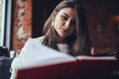 Kafeteryada oturan ve not defterindeki notları okuyan uzun saçlı serbest çalışan genç bir bayan.