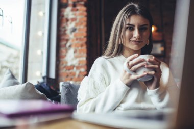 İyimser kadın uzaktan kumandalı beyaz süveter giymiş kafeteryada bir fincan kahveyle otururken kameraya bakıyor.