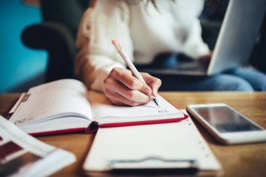 Tanımlanamayan kadın uzaktan çalışan sıradan giysiler içinde masada oturup proje üzerinde çalışırken planlayıcıya notlar yazan.