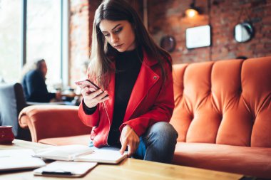 Kafedeki masada elinde defterle oturan konsantre kız öğrenci ve görev sırasında cep telefonunu mesajlaşmak için kullanıyor.