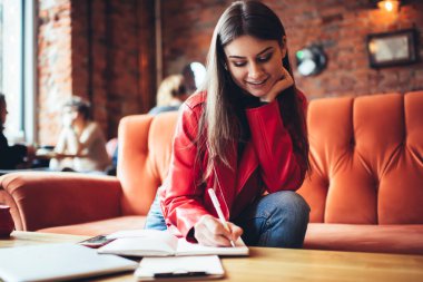 Neşeli bayan öğrenci, günlük kıyafetlerini giymiş, kanepeye yaslanmış kafeteryada ödev yaparken not alıyor.