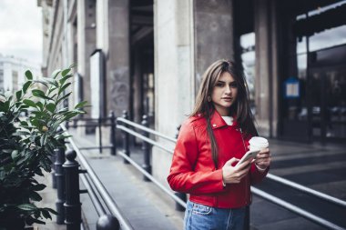 Uzun saçlı, sıradan giysili, hasretli bir kadın sokakta durup akıllı telefonlara bakarken kameraya bakıyor.