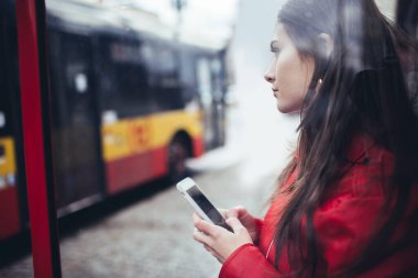 Otobüs durağındaki akıllı telefondan mesajlaşırken uzun saçlı, sıradan giyinmiş düşünceli bir kadının yan görüntüsü.