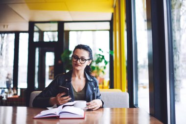 Gözlüklü, bir fincan lezzetli kahveyle masada oturan ve akıllı telefondan proje hakkında bilgi toplayan konsantre kadın serbest çalışan.