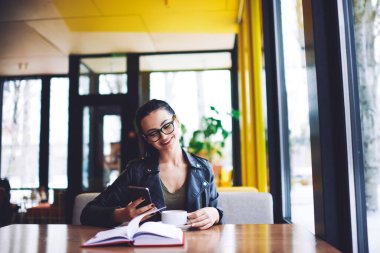 Kafenin penceresinin yanındaki masada oturup kahve içerken mutlu bir kadın cep telefonuna mesaj atıyor.