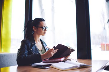 Kafe 'de kitap okurken pencerenin yanındaki masada oturan günlük kıyafetli kız öğrencinin yan görüntüsü.