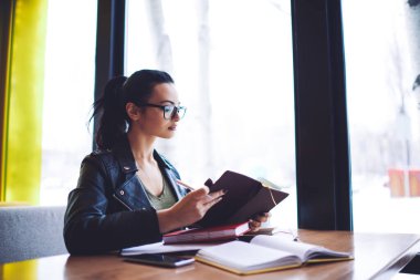 Kafe 'de kitap okurken pencerenin yanındaki masada oturan günlük kıyafetli kız öğrencinin yan görüntüsü.