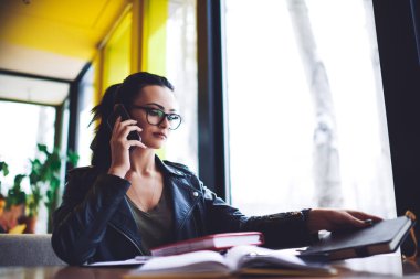 Düşük açılı, günlük kıyafetli, gözlüklü, pencerenin yanındaki masada oturan akıllı telefondan konuşan ve not defteri alan düşünceli bir kadın serbest yazar.