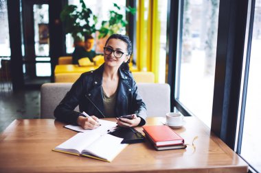 Kafede akıllı telefonlu bir masada otururken kameraya bakan ve not defterine not alan pozitif kadın serbest yazardan.