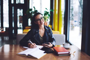 Genç, mutlu, gözlüklü, dişlek, gülümsemeli, cep telefonuyla masada oturmuş not defterine notlar yazarken kameraya bakan uzaktan kumandalı bir kadın.