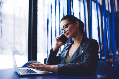 Gündelik kıyafetli, düşünceli bir kadın, pencerenin yanındaki masada oturmuş bilgisayarını karıştırıyor ve cep telefonuyla sohbet ediyor.