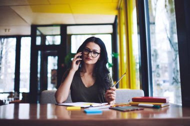 Düşünceli genç bayan girişimci ahşap masada kağıt ve kalemlerle otururken ve kafeteryada kalem taşırken cep telefonuyla iletişim kuruyor.