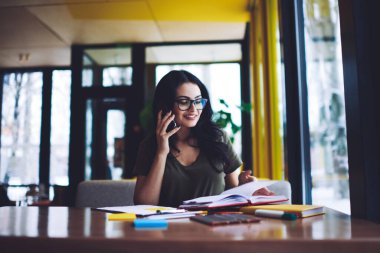 Modern kafeteryadaki kağıt ve kalemlerle masasında oturan, çağdaş akıllı telefon aracılığıyla konuşan ve kitaplarda notlar okuyan pozitif, genç bir esmer.