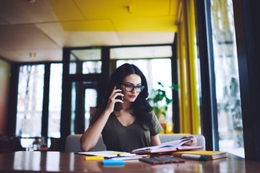 Odaklanmış koyu renk saçlı, günlük giysiler içinde genç bir kadın ahşap masada oturuyor ve belgelere bakarken cep telefonuyla konuşuyor.