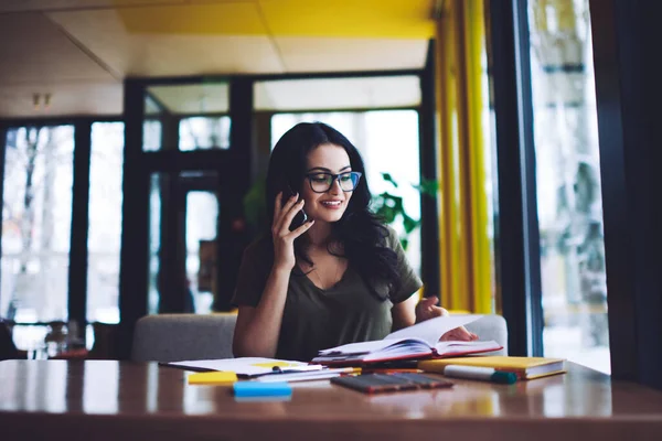 Modern kafeteryadaki kağıt ve kalemlerle masasında oturan, çağdaş akıllı telefon aracılığıyla konuşan ve kitaplarda notlar okuyan pozitif, genç bir esmer.