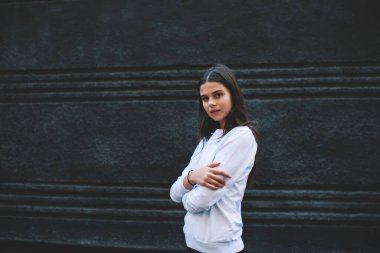 Dost canlısı, beyaz kazak ve bilezik giyen, kolları katlanmış, siyah arka planda duran ve kameraya bakan genç bir kadın.