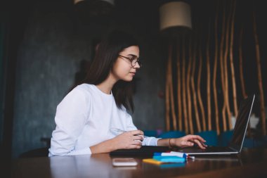 Masada oturmuş netbook ekranına bakan gözlüklü ve günlük giysileri olan konsantre bir bayan öğrencinin yan görüntüsü.
