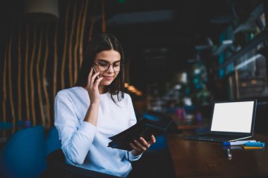 Mutlu kadın uzaktan kumandalı, dişlek gülüşlü, dizüstü bilgisayarla masada oturan ve telefon görüşmesi yaparken defteri açan kadın.