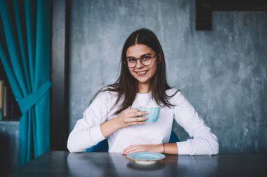 Tatmin olmuş bir kadın, dişlek bir gülümsemeyle masada çay tabağı ve bir fincan kahveyle kameraya bakarken.
