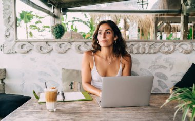 Kafeteryada bir bardak kahve ve gündemle masaya bakan genç, düşünceli, etnik bayan uzaktan kumandalı bir çalışan.