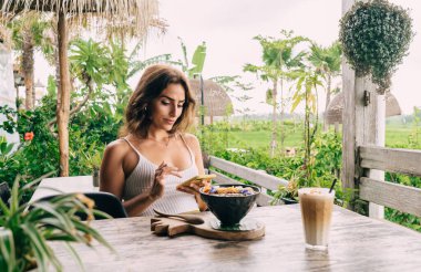 Moda kıyafetleri içinde kendinden emin genç bir kadın yaz kafesinde sağlıklı yemek kasesi ve buzlu kahveyle dinlenirken cep telefonuyla mesajlaşıyor.