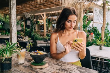 Genç etnik kadın cep telefonuyla mesajlaşırken yaz aylarında kafeterya masasında nefis kahve ve kahvaltı tabağıyla oturuyor.