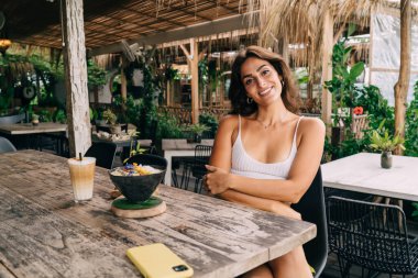 Kolları kavuşturulmuş gülümseyen etnik kadın, tropik tatil köyünde kahvaltı kasesi ve kahveyle masaya otururken kameraya bakıyor.