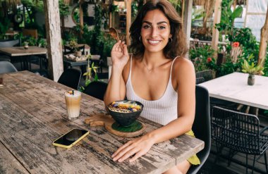 Dirsek dirseğinde eğilmiş, gülümseyen ve bir kase lezzetli yemek ve kahveyle masada otururken kameraya bakan mutlu etnik kadın.