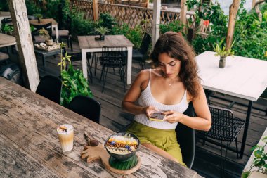 Kahve ve kahvaltı kâsesiyle servis masasında otururken cep telefonuyla mesajlaşan odaklanmış etnik kadın mesajlarından.