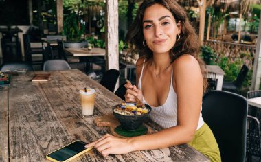 Günlük kıyafetleriyle neşeli pozitif kadın kahvaltıda lezzetli sağlıklı yiyecekler yiyor ve tropik tatil beldesindeki terasta otururken modern cep telefonlarını karıştırıyor.