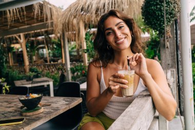 Açık hava restoranında oturan, tropik bir ülkede tatilini geçirirken soğuk ve lezzetli buzlu kahvenin tadını çıkaran ve gülümseyerek kameraya bakan beyaz elbiseli neşeli genç kadın.