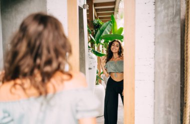 Modayı takip eden kıyafetleri içinde pozitif, şık bir kadın. Gülümseyen ve aynanın yanında duran yaz günündeki yansımaya neşeyle bakan.