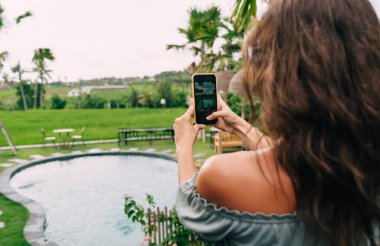 Yeşil tropikal bahçede gölün kenarında duran ve yaz günü cep telefonuyla fotoğraf çeken isimsiz bir kadının arka planı.