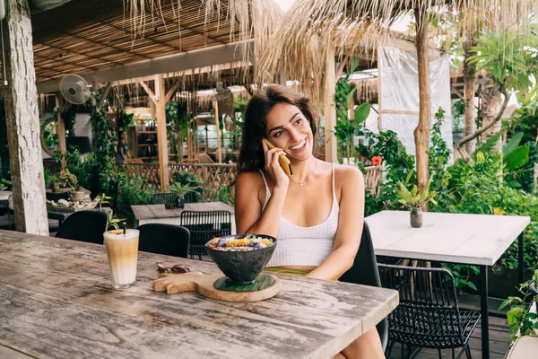熱帯リゾートの屋外カフェで朝食を食べながら携帯電話で話す白いトップを身に着けている若い魅力的な女性を笑顔 — ストック写真
