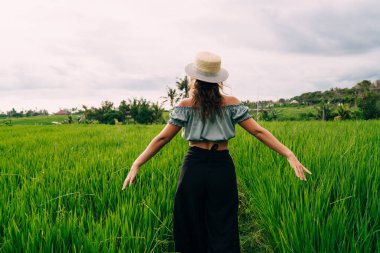 Şapkalı, uzun etekli, kolları ayrık bir şekilde Bali kırsalındaki yeşil patikada gezinen tanınmamış bir kadının arka görüntüsü.