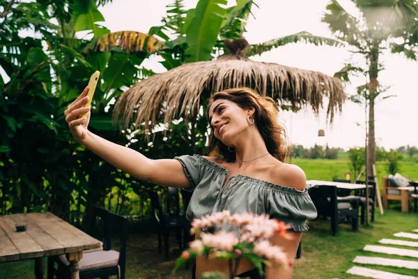 身着夏装 身材苗条的西班牙裔年轻女子站在巴厘岛热带旅游胜地花园时 一边笑 一边用智能手机自拍 — 图库照片