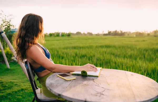田んぼを見つめ テキストのアイデアを考える女性作家の考え方 教育ノートやスマートフォンを持つ夏のテラスに座っている瞑想ブロガー — ストック写真