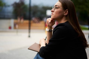 Görme düzeltmesi için optik gözlüklü düşünceli bayan öğrenci açık havada eğitimi düşünen, eskiz defteriyle şehirde oturan ve öğrenme ve öğrenme fikri üzerinde kafa yoran zeki kadın.